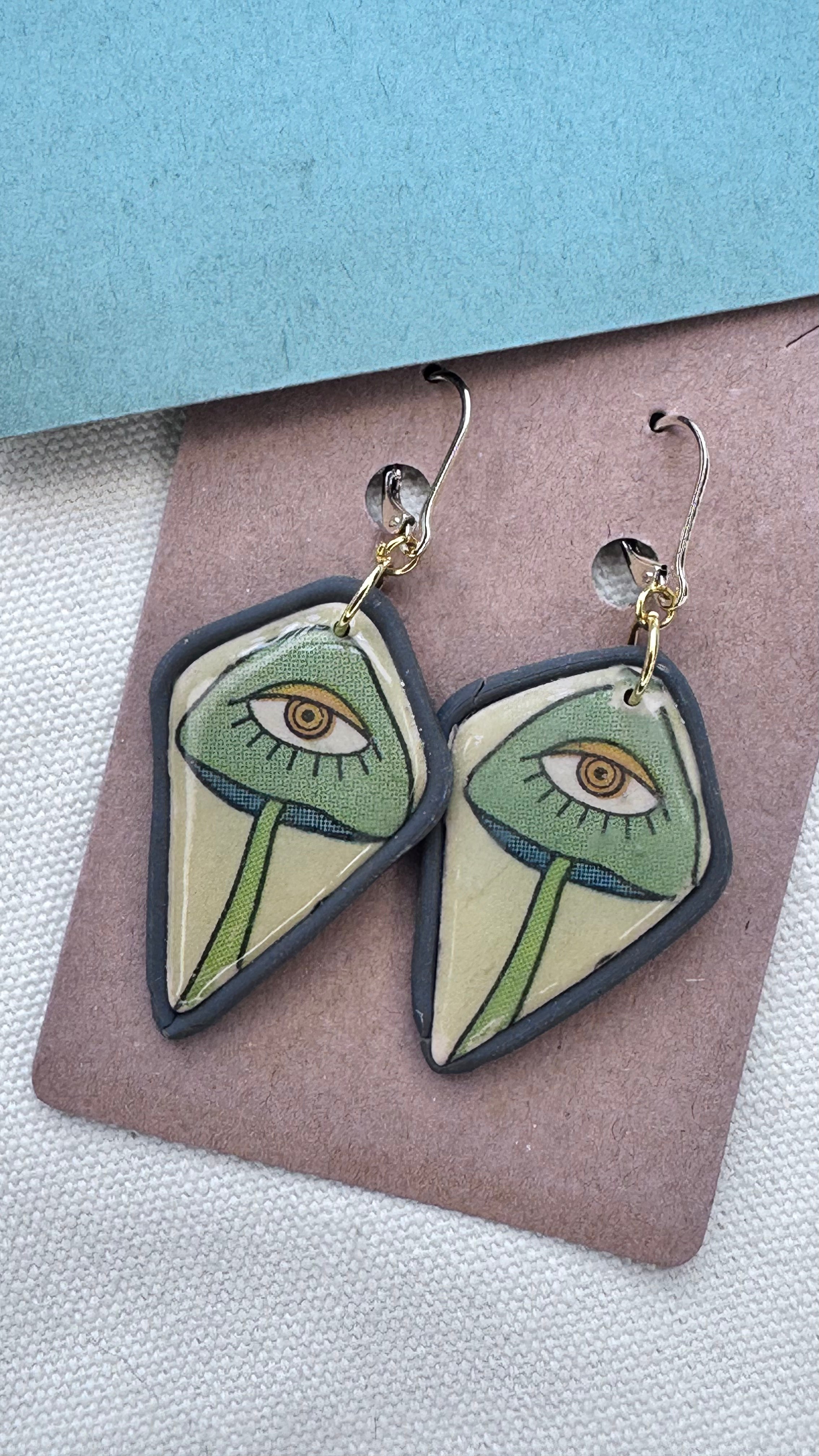 Dangle earrings featuring green eye illustrations on cream clay with charcoal gray borders and gold hooks, laid on kraft card with textured background