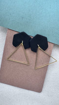 Black hexagon polymer clay earrings with brass triangle dangles on kraft backing, displayed against a blue and fabric background