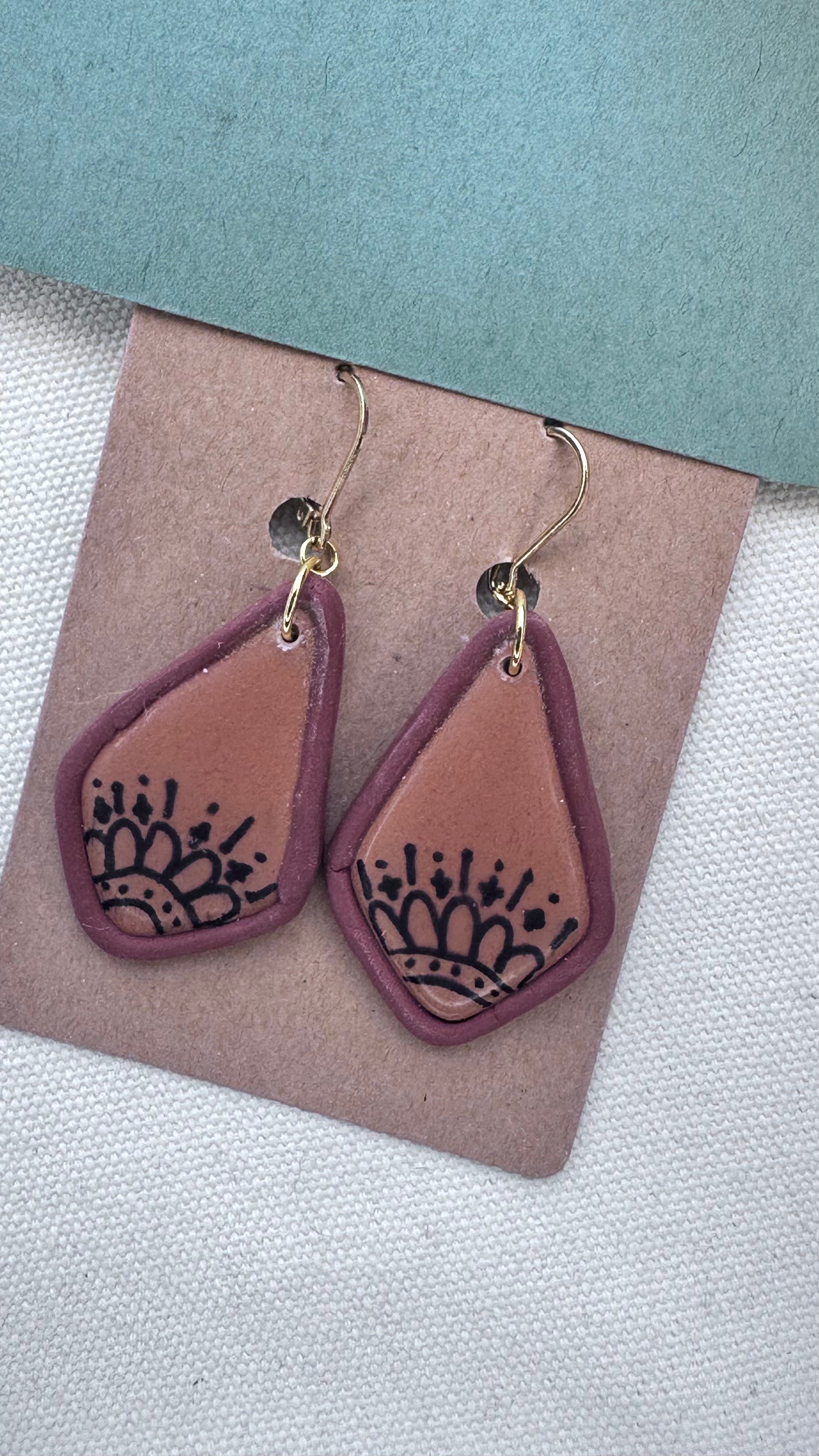 Terracotta and plum clay earrings with stamped black folk-inspired print, teardrop shape, and gold hooks on kraft backing