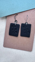 Matte black rectangular clay earrings with embossed botanical design and silver hooks, displayed on a kraft card background