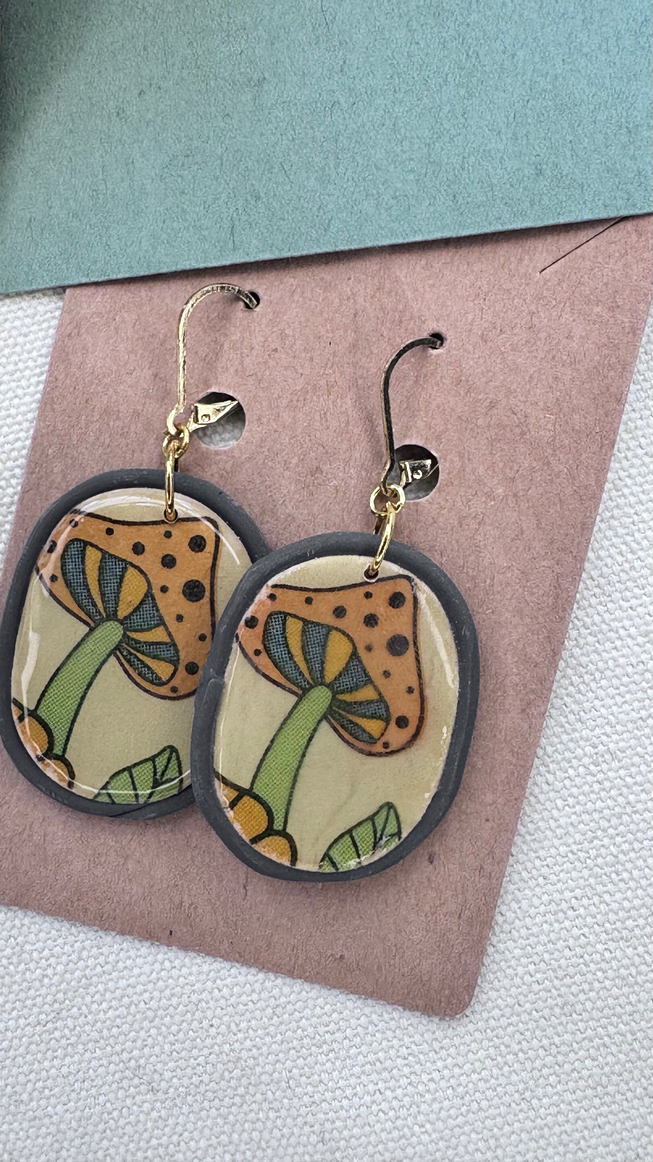 Oval resin-coated clay earrings with orange and green mushroom illustration, framed in dark gray with gold hooks, displayed on kraft card