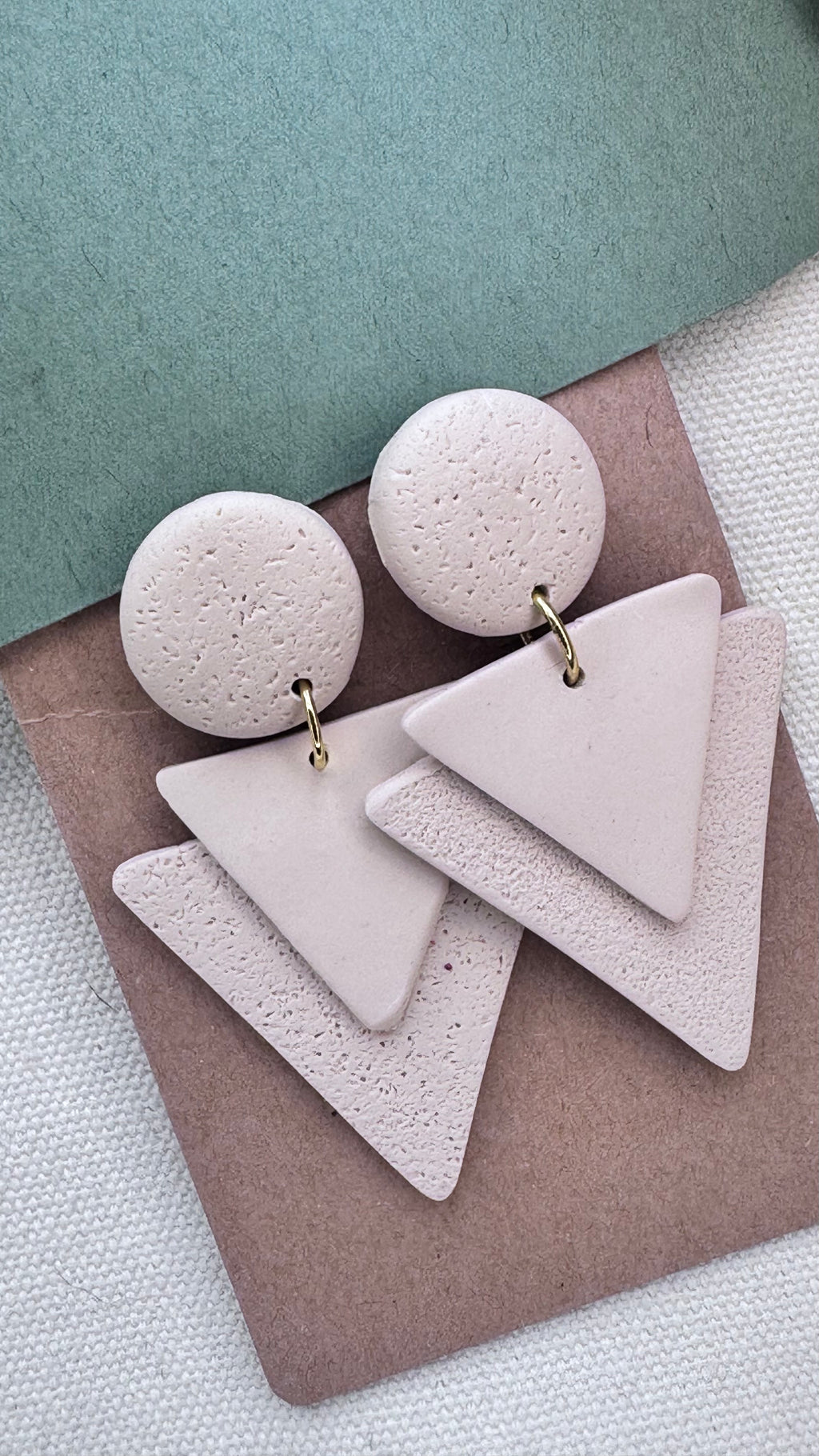 Modern earrings featuring a blush round top and textured triangle dangle made of polymer clay, laid on a kraft card over fabric