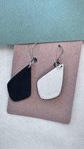 Black and white asymmetrical clay earrings with silver hooks, displayed on kraft card with blue and cream background